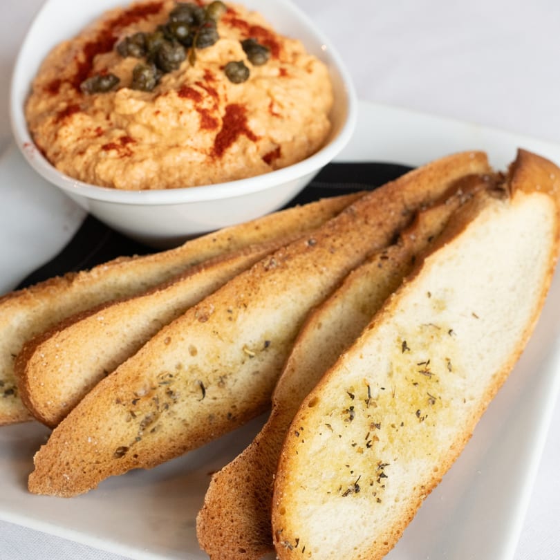 A white bowl of hummus topped with capers and paprika, served with toasted bread slices.